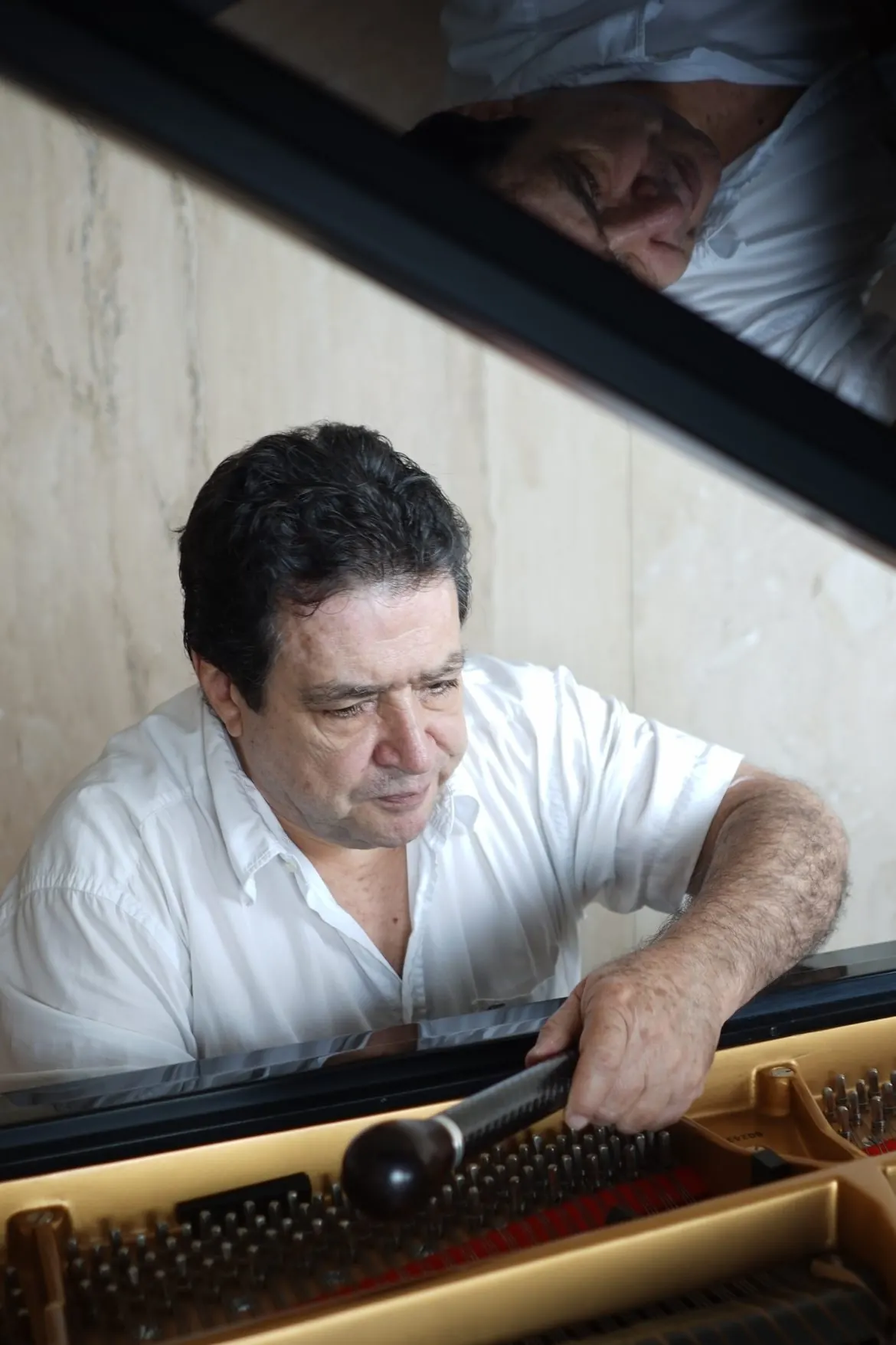 Rafael Melo afina un piano de cola con la llave de afinar; su reflejo aparece en la tapa abierta.